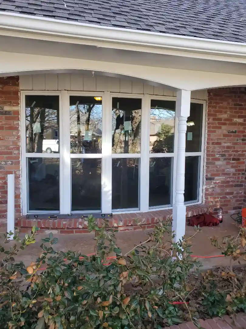 Newly installed large white-framed windows in a red-brick home with decorative exterior column and shrubbery in the foreground